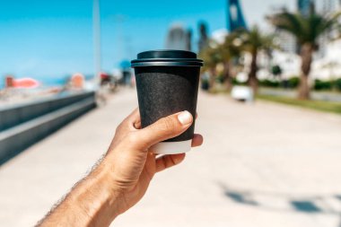 Bir el, sahil kentinde güneşli bir sahil bulvarının arka planında logosu olmayan siyah bir kahve fincanını tutuyor. Fotoğraf, şehir eğlencesi ve modern hayatın özünü yansıtıyor.