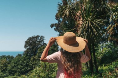 Açık pembe elbiseli ve hasır şapkalı genç bir kadın sırtı kameraya dönük, parlak mavi gökyüzünün altındaki yoğun yeşil ormanı seyrediyor. Kırmızımsı pembe uçlu uzun saçları sırtından aşağı akıyor.