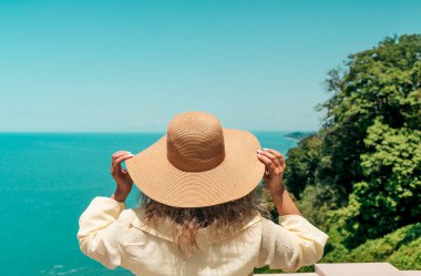 Hafif bir yaz kostümü ve geniş kenarlı hasır şapkalı bir kadın manzaralı bir uçurumun kenarında uçsuz bucaksız deniz ufkuna bakıyor. Duruşu huzur, yansıma ve seyahat arzusunu çağrıştırıyor. Parlak