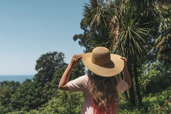 Açık pembe elbiseli ve hasır şapkalı genç bir kadın sırtı kameraya dönük, parlak mavi gökyüzünün altındaki yoğun yeşil ormanı seyrediyor. Kırmızımsı pembe uçlu uzun saçları sırtından aşağı akıyor.