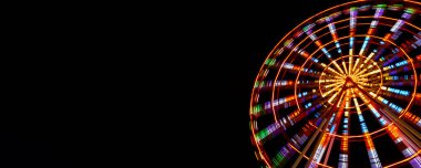 Göz kamaştırıcı neon ışıklarla aydınlatılmış canlı bir dönme dolap gece vakti bir lunaparkta derin siyah gökyüzüne doğru döner. Uzun pozlama hareketi yakalar ve hipnotik dairesel bir etki yaratır.