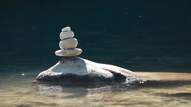 Dengeli bir taş yığını, aydınlık bir yaz gününde bir nehir kıyısında huzur içinde duruyor. Yumuşak su yakınlarda akar ve rahatlatıcı doğal bir zemin oluşturur. Bu rahatlatıcı sahne farkındalık hissini uyandırıyor.