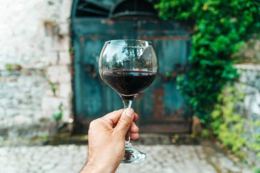 Geleneksel bir taş şarap mahzeninin önünde kırmızı şarap kadehini tutan bir elin yakın çekimi, Fransa ya da İtalya 'nın zengin şarap yapma mirasını anımsatıyor. Görüntü, eski dünya cazibesini taşır.