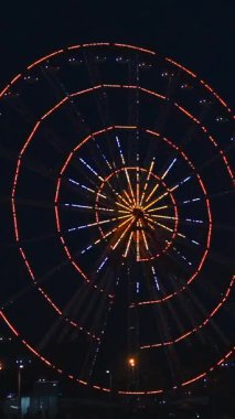 Göz kamaştırıcı neon ışıklarla aydınlatılmış canlı bir dönme dolap gece vakti bir lunaparkta derin siyah gökyüzüne doğru döner. Uzun pozlama hareketi yakalar ve hipnotik dairesel bir etki yaratır.