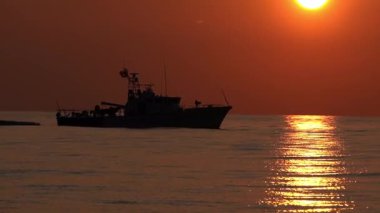 Deniz kurtarma botu dramatik bir akşam günbatımında sakin sularda süzülüyor. Geminin silueti parlayan gökyüzüne karşı duruyor. Uyanıklık, güvenlik ve acil durumu simgeliyor.
