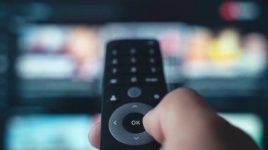 Uzaktan kumandayı tutan bir el, arka planda bulanık bir ekran olan televizyondaki kanalları değiştirir. Görüntüler modern medya tüketimini, içerik aşırı yüklemesini ve dijital seçim yorgunluğunu simgeliyor.