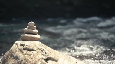 Dengeli bir taş yığını, aydınlık bir yaz gününde bir nehir kıyısında huzur içinde duruyor. Yumuşak su yakınlarda akar ve rahatlatıcı doğal bir zemin oluşturur. Bu rahatlatıcı sahne farkındalık hissini uyandırıyor.
