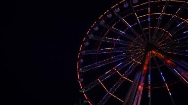 Göz kamaştırıcı neon ışıklarla aydınlatılmış canlı bir dönme dolap gece vakti bir lunaparkta derin siyah gökyüzüne doğru döner. Uzun pozlama hareketi yakalar ve hipnotik dairesel bir etki yaratır.