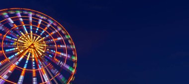 Göz kamaştırıcı neon ışıklarla aydınlatılmış canlı bir dönme dolap gece vakti bir lunaparkta derin siyah gökyüzüne doğru döner. Uzun pozlama hareketi yakalar ve hipnotik dairesel bir etki yaratır.