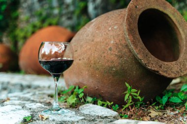 Geleneksel Gürcistan qvevri 'sinin yanında bir bardak kırmızı şarap duruyor. Eski şarap yapımında kullanılan büyük kil kaplar. Kırsal bir ortamda geçen sahne, Gürcistan bağcılık ve doğal fermantasyon yöntemlerinin zengin mirasını vurguluyor. Bu resim... 