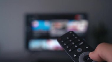 Uzaktan kumandayı tutan bir el, arka planda bulanık bir ekran olan televizyondaki kanalları değiştirir. Görüntüler modern medya tüketimini, içerik aşırı yüklemesini ve dijital seçim yorgunluğunu simgeliyor.