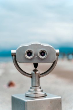 Arka planda bulanık bir kıyı manzarası olan çakıl taşı plajında paslanmaz çelik bir dürbün görüntüleyicisinin yakın çekimi. Görüntü deniz kenarındaki turizm, keşif ve yaz tatillerini çağrıştırıyor.