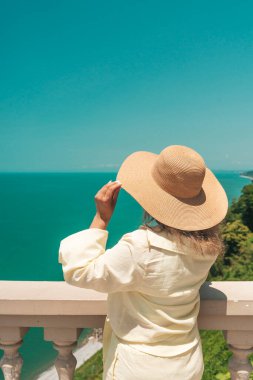Hafif bir yaz kostümü ve geniş kenarlı hasır şapkalı bir kadın manzaralı bir uçurumun kenarında uçsuz bucaksız deniz ufkuna bakıyor. Duruşu huzur, yansıma ve seyahat arzusunu çağrıştırıyor. Parlak