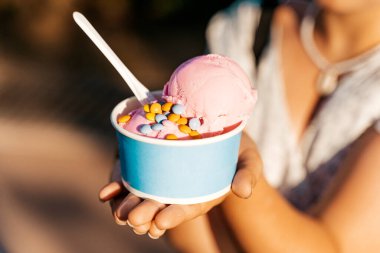 Pembe çilekli dondurma ve açık havada güneşli bir park ortamında tutulan renkli şekerlerle dolu bir kağıt bardak. Bu neşeli yaz ziyafeti neşeyi, rahatlığı ve basit zevkleri anımsatıyor.