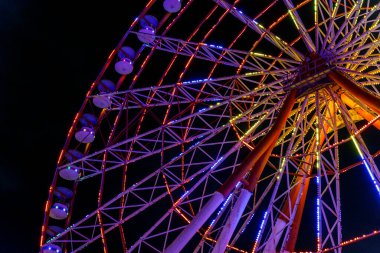 Göz kamaştırıcı neon ışıklarla aydınlatılmış canlı bir dönme dolap gece vakti bir lunaparkta derin siyah gökyüzüne doğru döner. Uzun pozlama hareketi yakalar ve hipnotik dairesel bir etki yaratır.