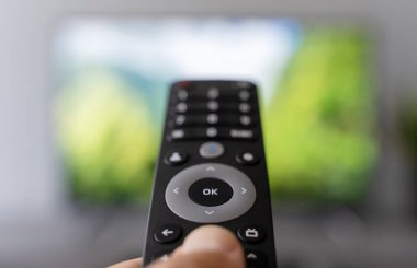 Uzaktan kumandayı tutan bir el, arka planda bulanık bir ekran olan televizyondaki kanalları değiştirir. Görüntüler modern medya tüketimini, içerik aşırı yüklemesini ve dijital seçim yorgunluğunu simgeliyor.