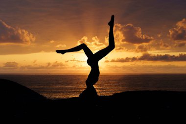 Hareketli bir günbatımında sakin bir sahil kenarında yoga yapan bir kadının huzurlu silueti. Bu sakinleştirici görüntü farkındalığı, iç dengeyi, ruhsal gelişimi ve bağlantıyı sembolize eder.