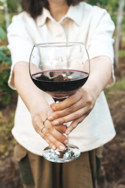 Bir kadının iki eli de yemyeşil üzüm bağında büyük bir bardak kırmızı şarap ikram etmesine yakın çekim. Görüntü, misafirperverlik, gelenek ve kırsal çekiciliği çağrıştırıyor.