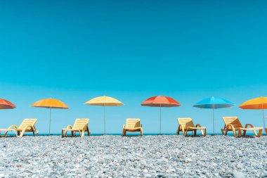 Parlak sarı güneş şezlongları ve çok renkli plaj şemsiyeleri açık mavi bir gökyüzünün altında çakıl taşı plajında mükemmel bir sırada dizilmiş. Görüntü rahatlatıcı tatil havasını yakalıyor, minimum yaz mevsiminde.