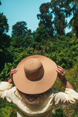 Açık sarı gömlekli ve geniş kenarlı hasır şapkalı bir kadının güneşli bir günde yemyeşil bir tropikal ormanın önünde dikilirken görüntüsü. Elleri nazikçe şapkayı tutuyor yeşile bakarken
