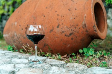 Geleneksel Gürcistan qvevri 'sinin yanında bir bardak kırmızı şarap duruyor. Eski şarap yapımında kullanılan büyük kil kaplar. Kırsal bir ortamda geçen sahne, Gürcistan 'ın zengin mirasını vurguluyor