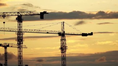 Gün batımında parlayan gökyüzüne çarpıcı bir şekilde kurulmuş inşaat vinçlerinin siluetleri. Bu görsel, kentsel gelişimin, mimari tutkuların ve