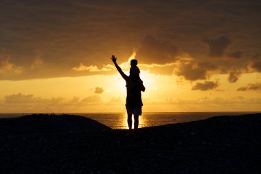 Baba ve oğul, gün batımının altın parıltısına karşı gölgede. Bu görüntü, ebeveyn ve çocuk arasındaki sevgi, bağlantı ve paylaşılan zamanın güzelliğini sembolize eden şefkatli ve neşeli bir anı yakalar.