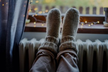 Women's legs feet in woolen socks on a heating radiator. Heating in winter concept, women's legs feet in woolen socks on a heating battery