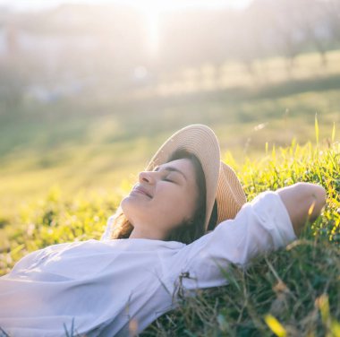 Dreamy young beautiful woman in hat lying on green grass with closed eyes enjoying summer sun and spring day
