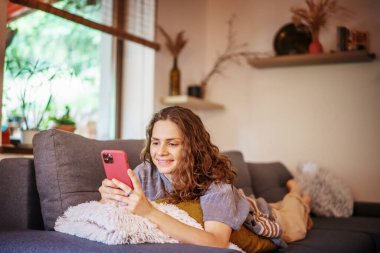 Cheerful smiling happy curly beautiful young woman lying relaxing on the sofa at home with a smartphone