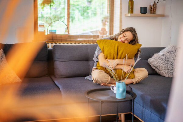 Cheerful curly cute young woman sitting at home on gray sofa hugging yellow pillow