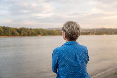 Elderly woman standing with her back on the river bank enjoying the view 