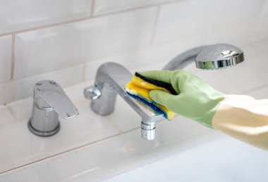 Cleaning in the bathroom. Woman's hand in a rubber glove close-up washing the faucet using a sponge. Cleanliness and hygiene concept