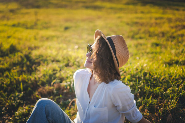 Молодая кавказская красивая женщина в белой рубашке соломенной шляпе солнцезащитные очки наслаждаясь весенним солнцем сидя на зеленой траве