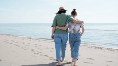 Genç lezbiyen çift deniz kıyısında çıplak ayakla birbirlerini kucaklayarak yürüyorlar.