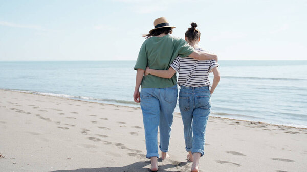 Молодая лесбийская пара, идущая босиком, обнимает друг друга за талию вдоль побережья
