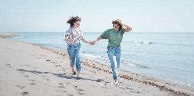 Genç, romantik lezbiyen çift deniz kıyısında el ele tutuşarak yalınayak koşuyor..
