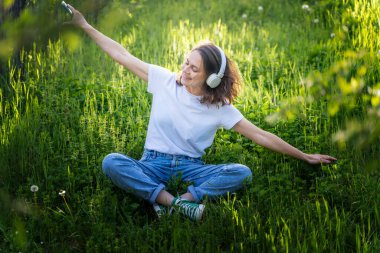 Genç, neşeli, rüya gibi bir kadın yaz bahçesinde oturmuş akıllı telefon ve kulaklık kullanarak müzik dinliyor.,