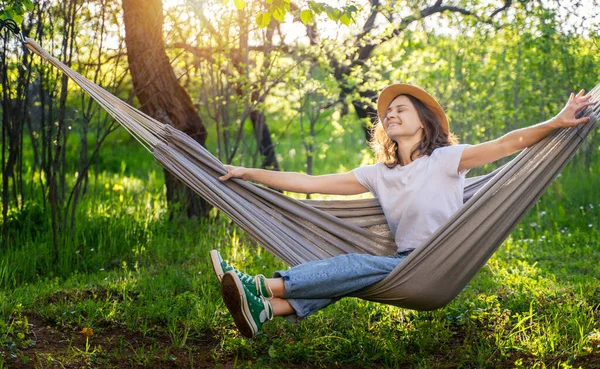 夏を楽しむ緑の庭でハンモックに横たわっている帽子の若い幸せな白人女性 — ストック写真