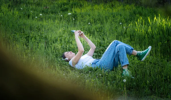 庭の緑の芝生の上に横たわる白いTシャツの若い陽気な女性は スマートフォンの画面を見て — ストック写真