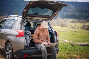 Orta yaşlı, beyaz bir adam açık araba bagajında oturuyor. Dağlık kırlarda dolaşırken dizüstü bilgisayar kullanıyor.