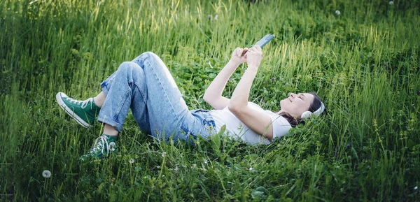 穿着白色T恤的年轻而快乐的女人躺在花园的绿草上 凝视着智能手机屏幕 — 图库照片