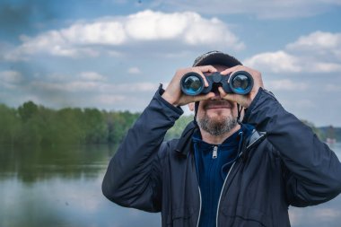 Beyaz, orta yaşlı bir adam vahşi doğada yürürken dürbünle bakıyor.