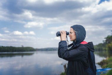Beyaz, orta yaşlı kadın nehir kıyısında yürürken dürbünle bakıyor.