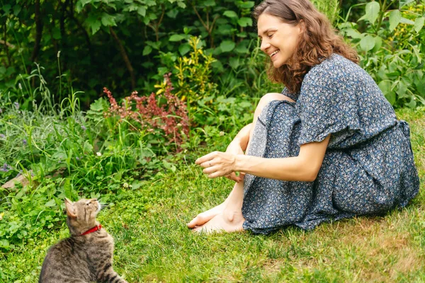 夏のドレスの若い陽気な笑顔の女性は 夏の庭で草の上に座っている灰色の猫と遊ぶ — ストック写真