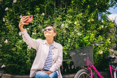 Çekici, mutlu, yaşlı bir kadın şehirde akıllı telefon kullanarak selfie çekiyor.