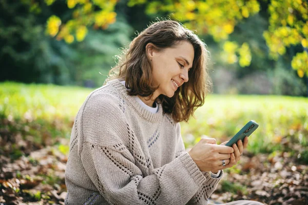 Очаровательная Кудрявая Молодая Женщина Сидит Осеннем Парке Мобильным Телефоном Руках — стоковое фото