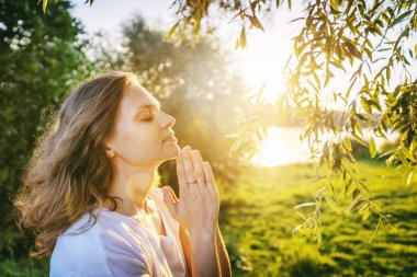 Gün batımında nehir kıyısında gözleri kapalı dua eden genç bir kadın.