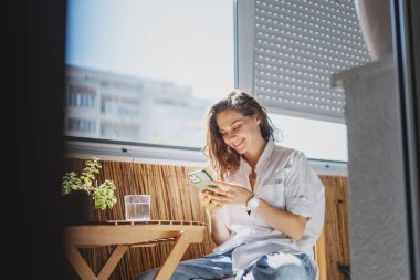 Masanın balkonunda oturan beyaz tişörtlü neşeli genç kadın akıllı telefonuyla.