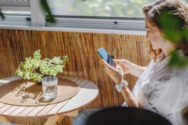 Güzel genç bir kadın apartmanın balkonunda oturmuş akıllı telefon kullanıyor.